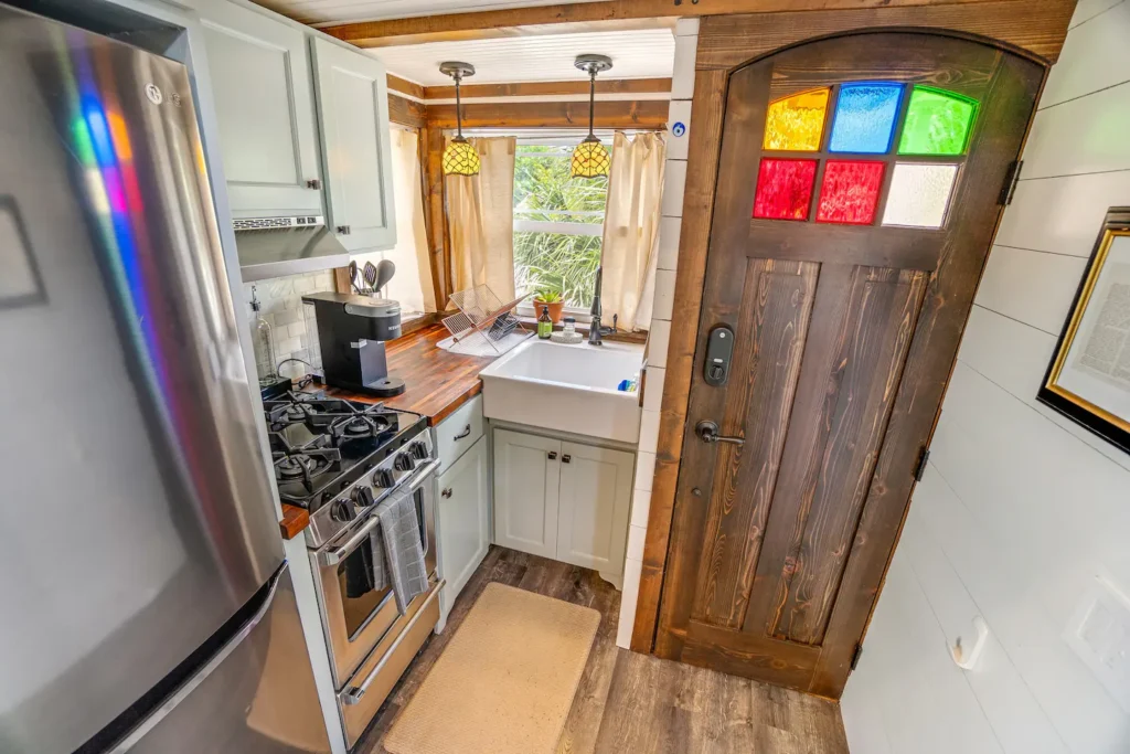 Kitchen in Tiffany The Tiny Home with a stainless steel refrigerator, gas range, white cabinets, farmhouse sink, and a wooden front door with stained glass panels