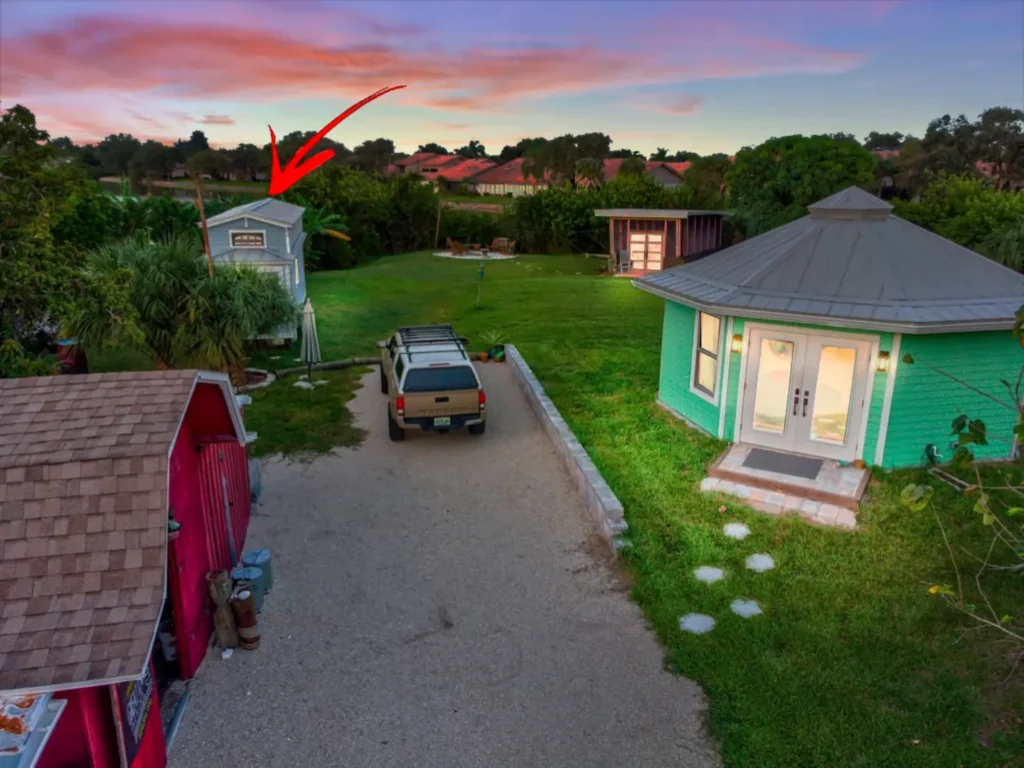 Aerial view of the shell driveway on Shellmate Island at sunset leading past the red shed toward Stanley the mint-green octagonal tiny house, with Tiffany The Tiny Home marked by a red arrow in the background