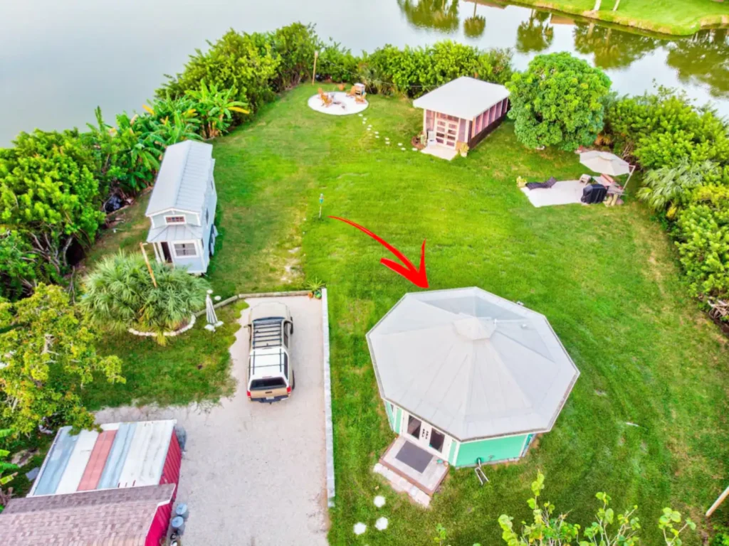 Aerial view of Shellmate Island in Sarasota, Florida, with Stanley the mint-green octagonal tiny house marked by a red arrow, Tiffany The Tiny Home on wheels, the fire pit and screened lounge, and water on both sides