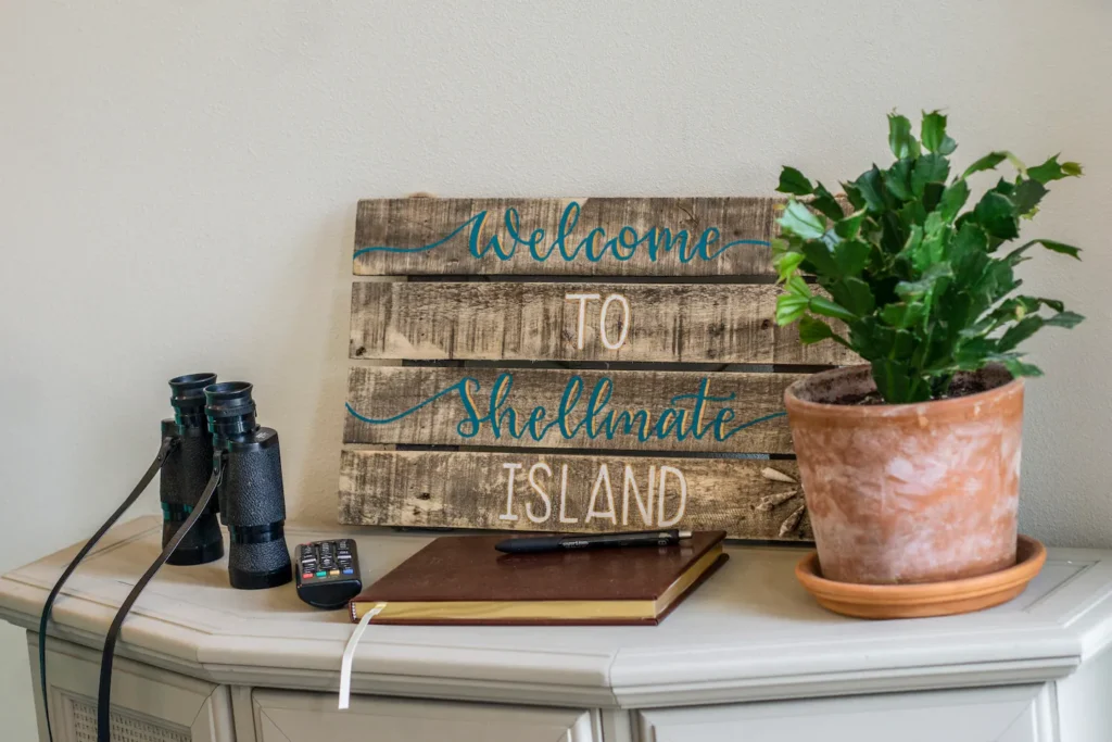 Rustic wooden Welcome to Shellmate Island sign on a dresser with binoculars, a guest book and a potted plant