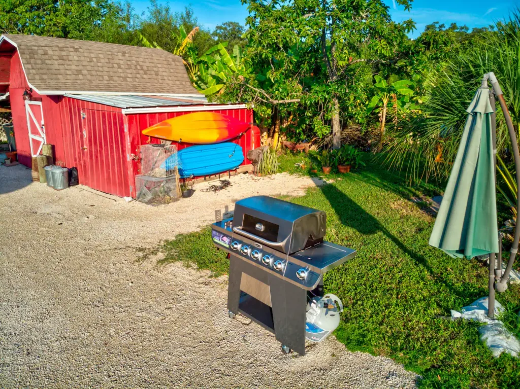 Outdoor cooking area on Shellmate Island with a gas grill, patio umbrella and a red storage shed holding kayaks and paddleboards, on shell gravel under banana plants