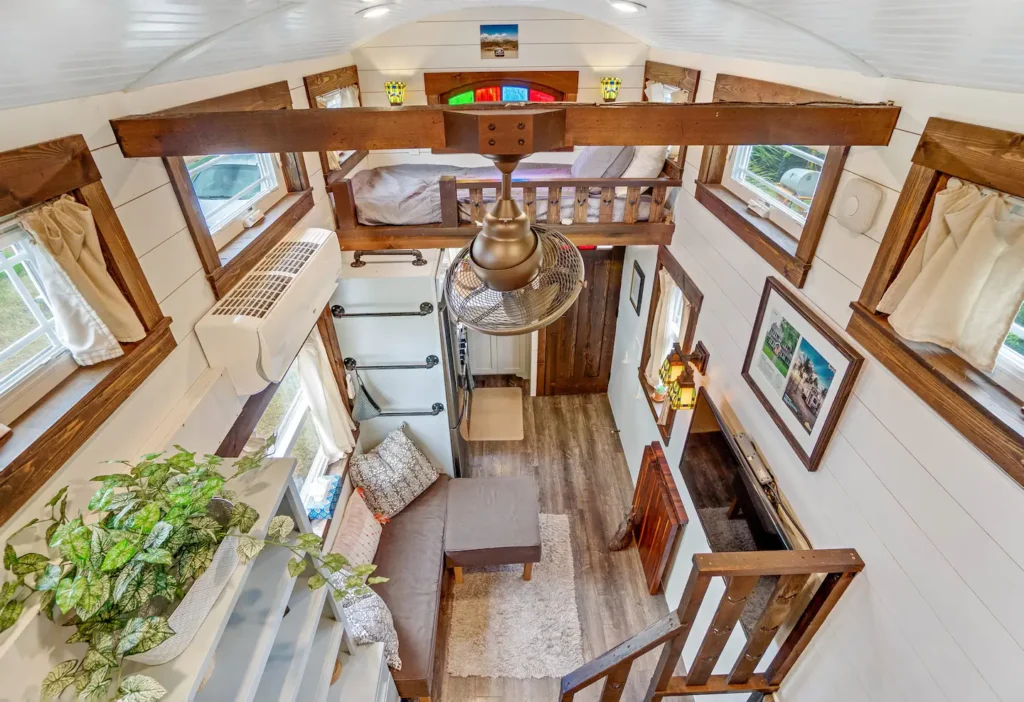 View from the sleeping loft of Tiffany The Tiny Home looking down into the living area with wood beams, a chandelier, built-in shelves, and windows on three sides