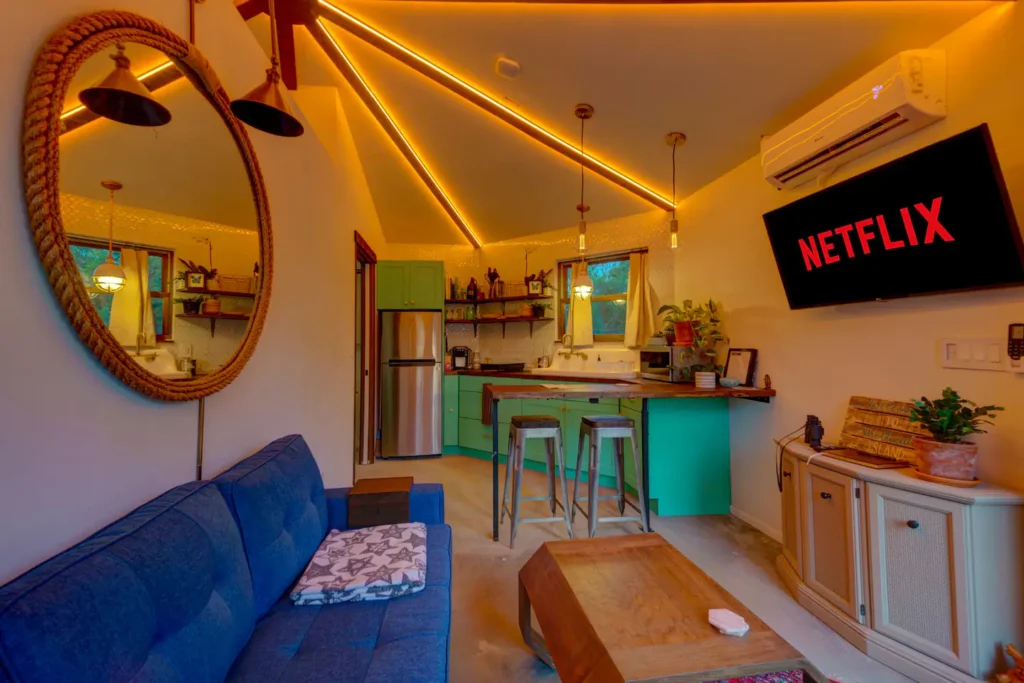 Living room in Stanley on Shellmate Island with warm amber LED lighting along the octagonal ceiling beams, a blue sofa, round rope mirror, wooden coffee table and the mint-green kitchen with bar stools