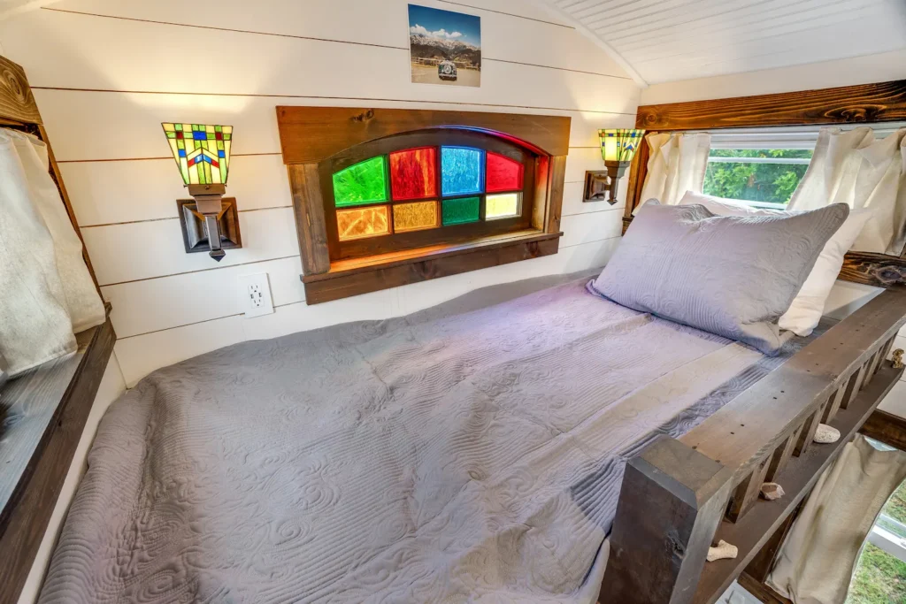 Sleeping loft in Tiffany The Tiny Home with a queen bed, stained glass window, wood beams, and white shiplap ceiling