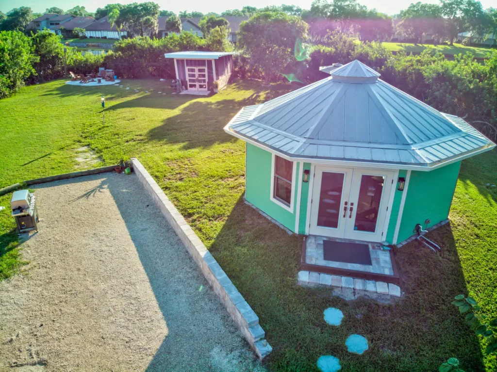 Aerial view of Stanley, the mint-green octagonal tiny house with a metal roof and red-trimmed French doors, on the lawn of Shellmate Island with the shell path and water behind