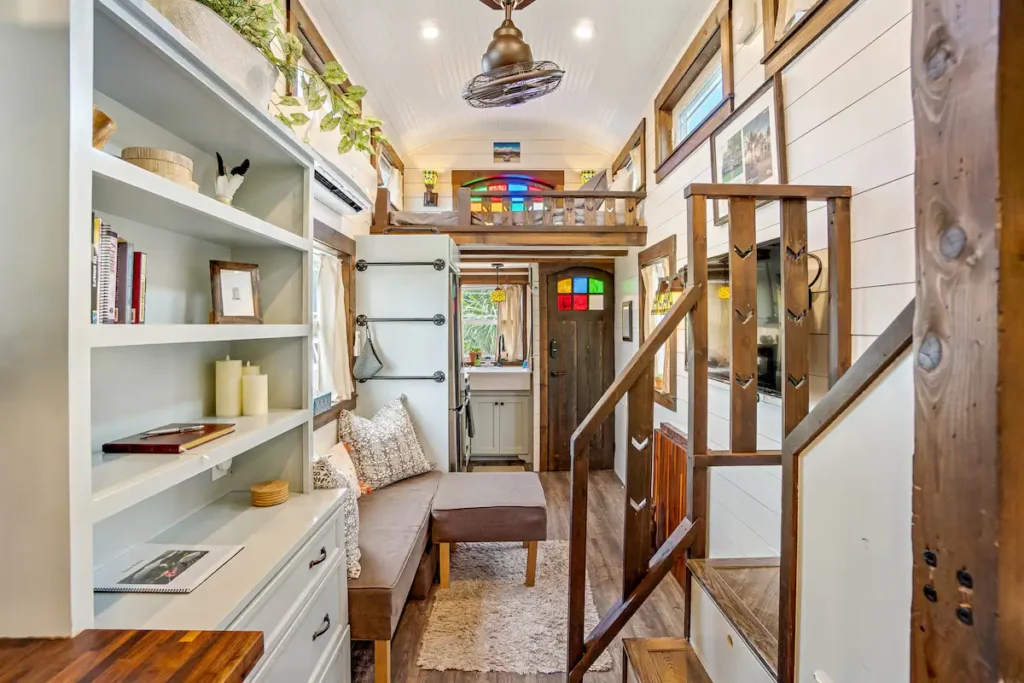 Living room of Tiffany The Tiny Home with built-in white shelving, a bench seat, mini-split air conditioner, stained glass accents, and the wooden staircase to the loft