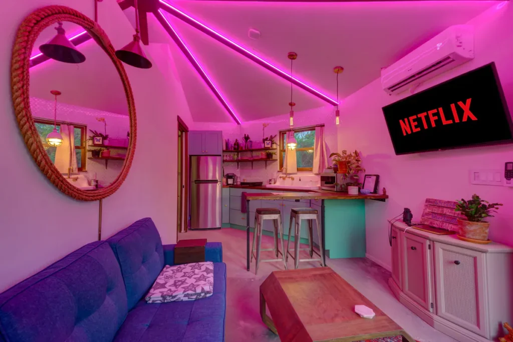 Interior of Stanley on Shellmate Island lit with pink and purple LED ceiling lighting, showing the blue sofa, round mirror, kitchen bar and Netflix on the TV