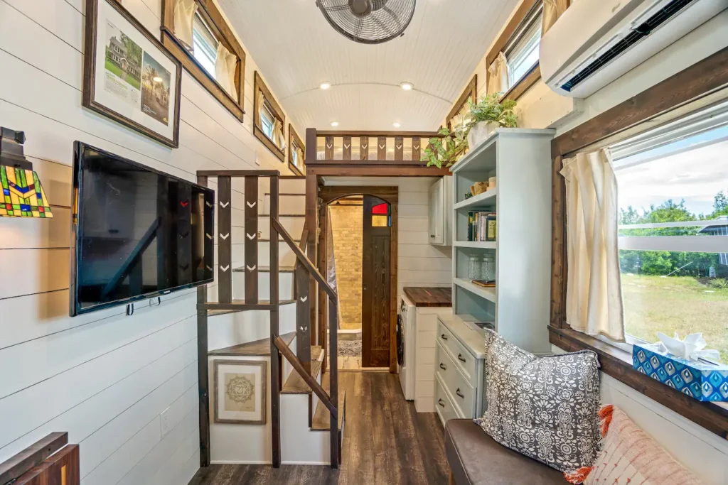 Living area of Tiffany The Tiny Home with a wall-mounted smart TV, white shiplap walls, storage stairs up to the sleeping loft, and a window facing the water