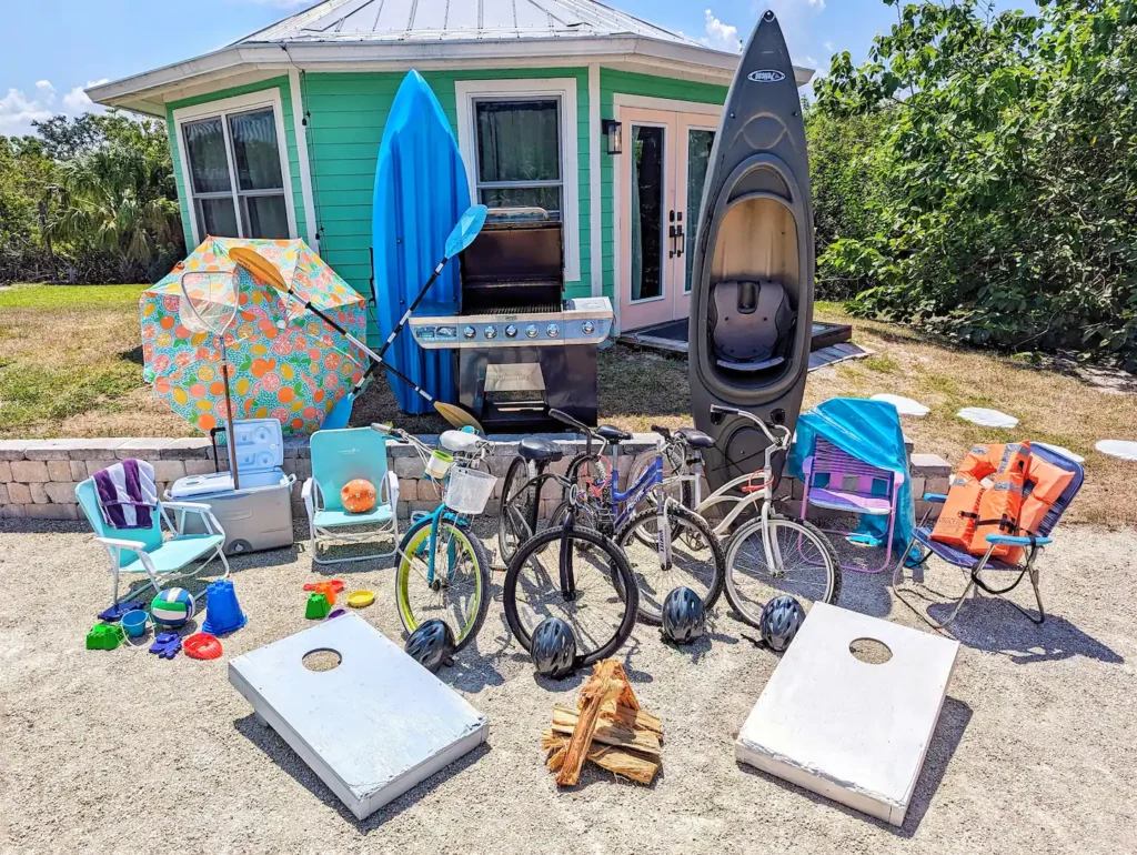 Free guest gear laid out in front of Stanley the mint-green tiny house on Shellmate Island: bicycles, kayaks, paddleboard, grill, cornhole boards, beach chairs, umbrella and games