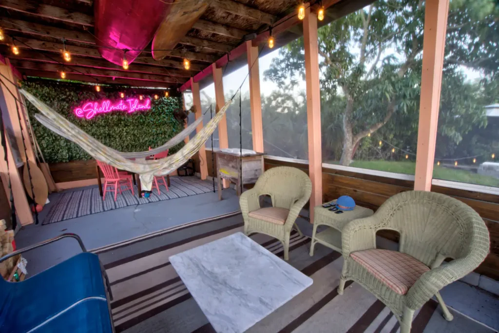 Screened-in lounge on Shellmate Island at night with string lights, a hammock, wicker chairs, blue couch, and the pink Shellmate Island neon sign