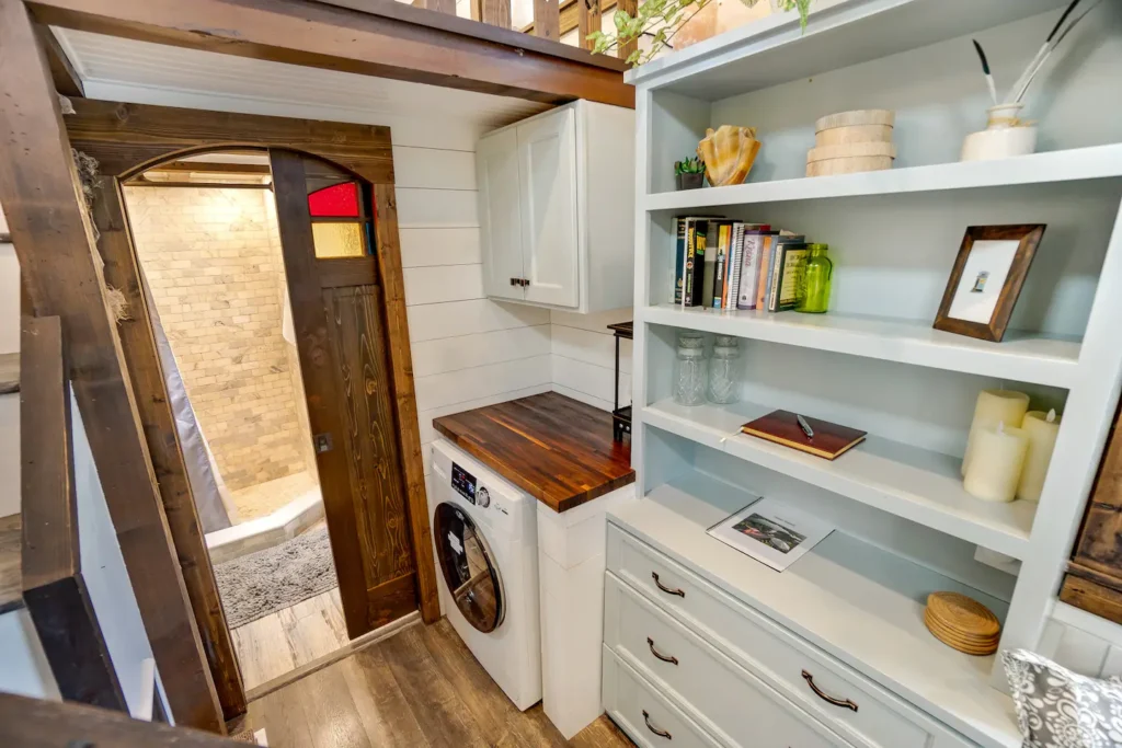 Interior of Tiffany The Tiny Home in Sarasota, Florida: white open kitchen shelving, built-in washer and dryer, and wood-trimmed doorway to the bathroom in a 270 sq ft tiny house on wheels