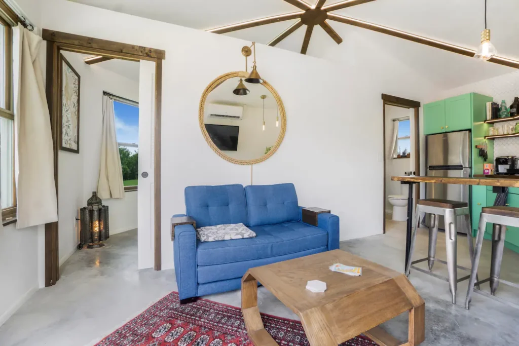 Living room in Stanley, the octagonal tiny house on Shellmate Island, with a blue sofa, wooden coffee table, round rope mirror, ceiling fan, red rug and a view toward the mint-green kitchen