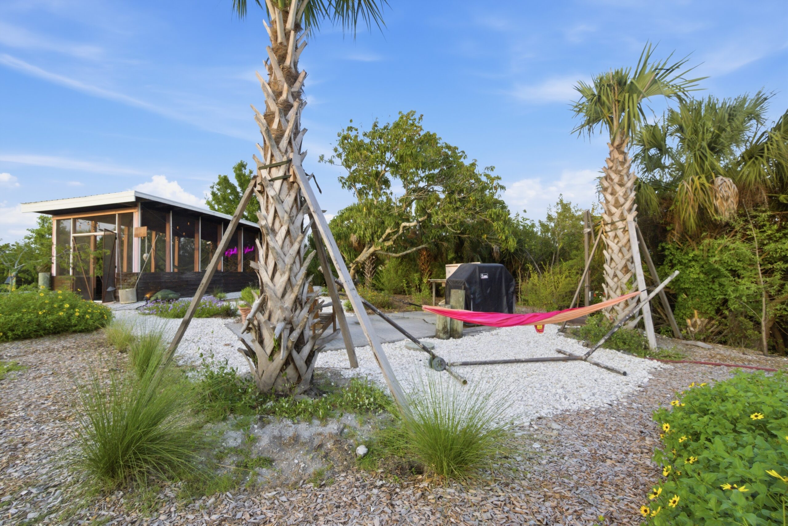 Shellmate Island tiny house retreat in Sarasota, Florida — screened-in communal lounge with pink hammock between sabal palms, white shell gravel pathways, and native butterfly garden plantings including beach sunflowers and muhly grass.