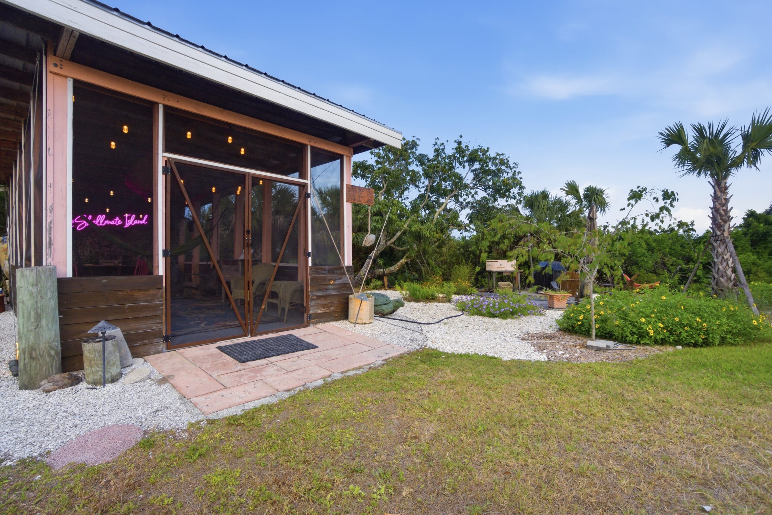 Shellmate Island tiny house retreat in Sarasota, Florida — screened-in communal lounge with pink hammock between sabal palms, white shell gravel pathways, and native butterfly garden plantings including beach sunflowers and muhly grass.