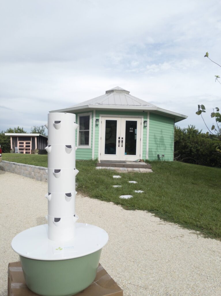 A white vertical tower garden planted with greens in front of the mint-green Shellmate Island tiny home on an overcast day