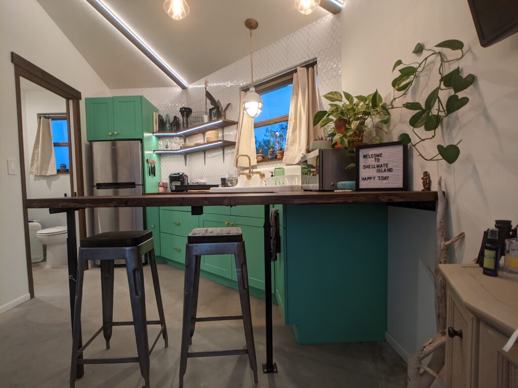 Teal kitchen with a wood breakfast bar and two black stools inside Shellmate Island tiny home, with a window view over the island