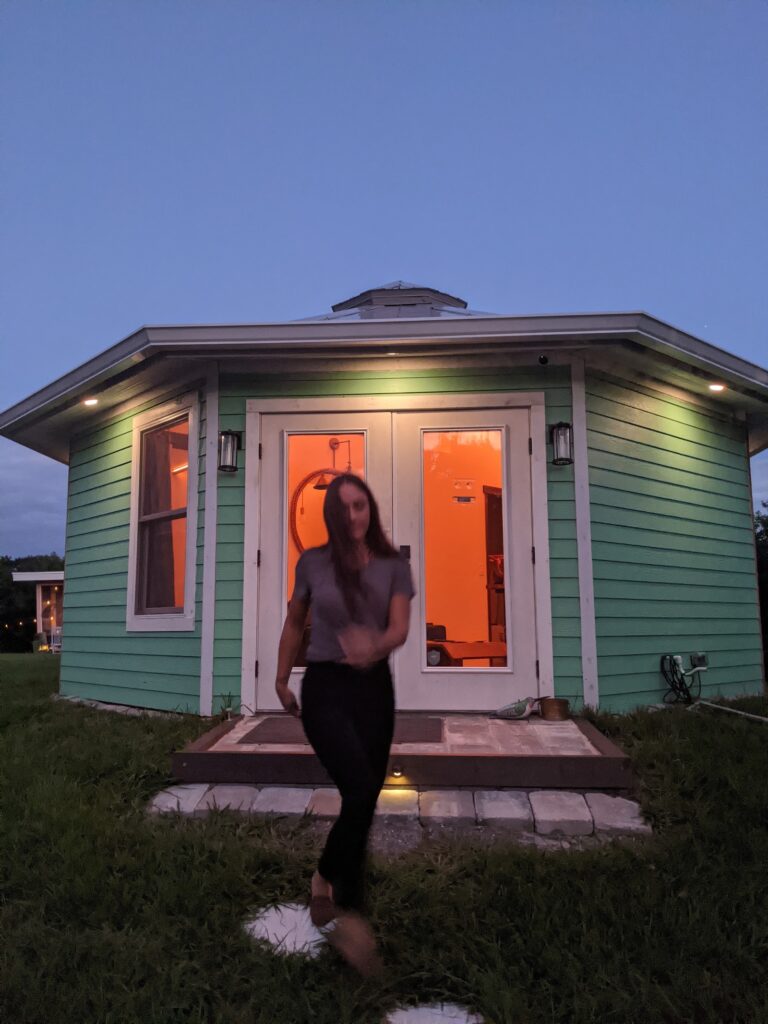 Shellmate Island tiny home at dusk with warm light glowing through the French doors and a guest standing on the path outside
