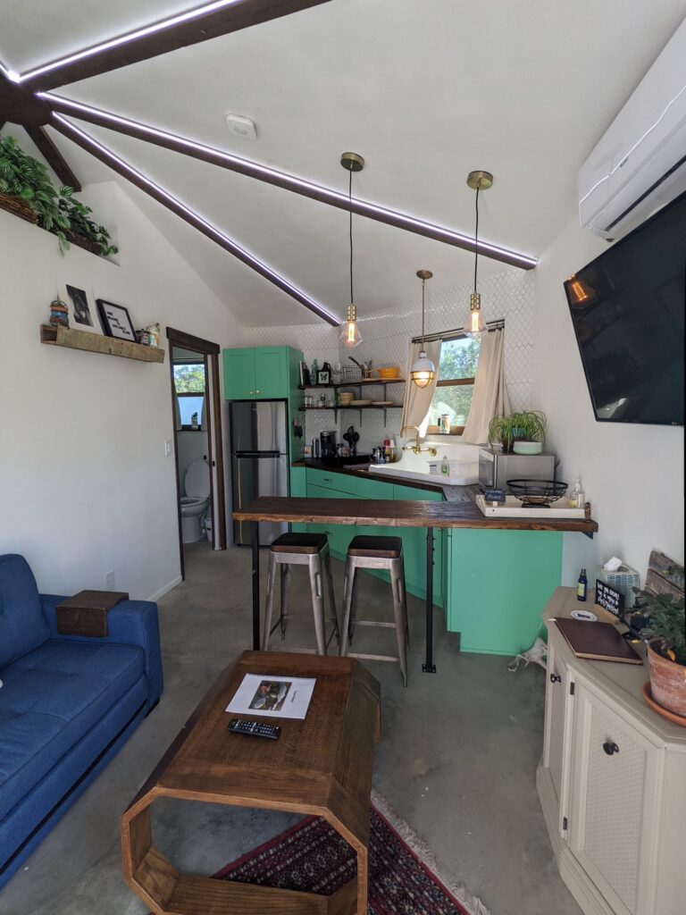 Inside Shellmate Island tiny home: blue sofa, teal kitchen cabinets, breakfast bar with stools, and the octagonal vaulted ceiling