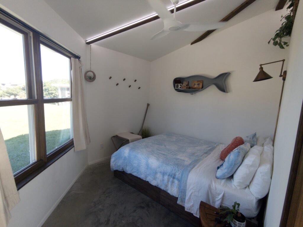 Bright bedroom inside Shellmate Island tiny home with a made bed, white walls, a fish wall ornament and a window looking out to the island